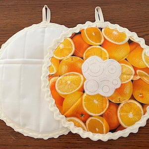 May include: Two white cotton pot holders with a scalloped edge. One pot holder has a white quilted design and the other has a print of orange slices.