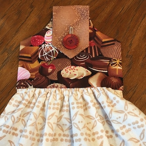 May include: A white tea towel with a brown and white patterned design. The top of the towel has a brown fabric pocket with a red button that says "Ragwear". The pocket is decorated with a chocolate and candy print.