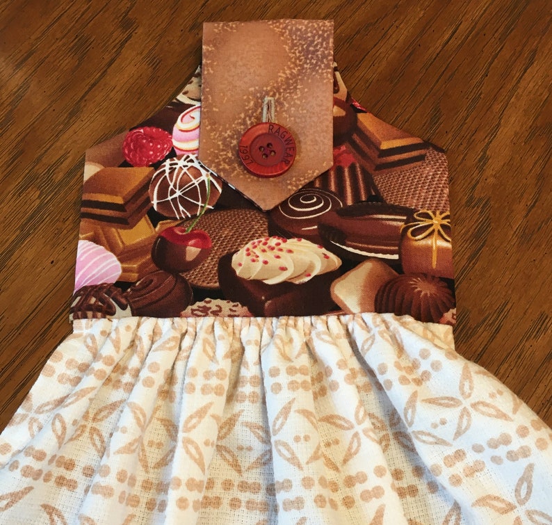 May include: A white tea towel with a brown and white patterned design. The top of the towel has a brown fabric pocket with a red button that says "Ragwear". The pocket is decorated with a chocolate and candy print.