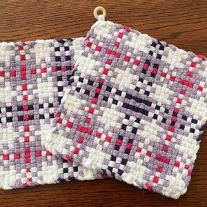 May include: Two woven pot holders with a checkered pattern in white, purple, pink, and black. The pot holders have wooden rings attached to the top corners.