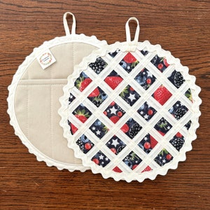 May include: Two round pot holders. One is beige with a white lace trim and the other is white with a white lace trim and a pattern of red strawberries, blackberries, and blueberries on a dark blue background.