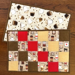 May include: A brown quilted placemat with a patchwork design featuring various coffee cup and beverage patterns. The placemat is made of fabric squares in brown, beige, red, and white with a coffee theme.