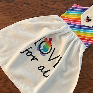 May include: White kitchen towel with a rainbow stripe border and a hanging loop. The towel has a black and white graphic of Mickey Mouse with the words "LOVE for all" printed on it.