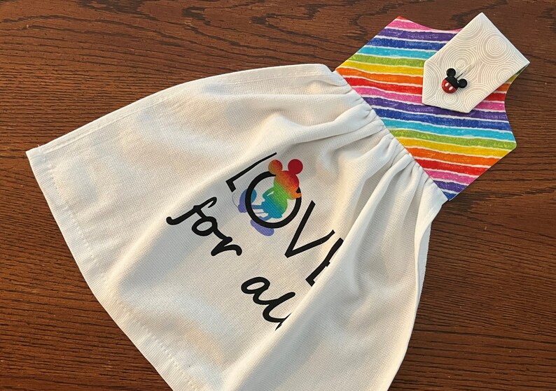May include: White kitchen towel with a rainbow stripe border and a hanging loop. The towel has a black and white graphic of Mickey Mouse with the words "LOVE for all" printed on it.