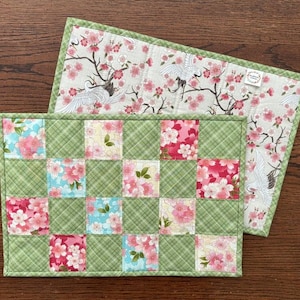 May include: A green and white patchwork placemat with a floral design. The placemat is made of squares of fabric sewn together. The placemat is 12 inches by 18 inches.