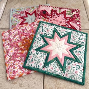 May include: Four handmade quilted pot holders with starburst designs. Each pot holder features a different floral pattern in shades of pink, red, and green. The pot holders are square, approximately 8 inches by 8 inches.