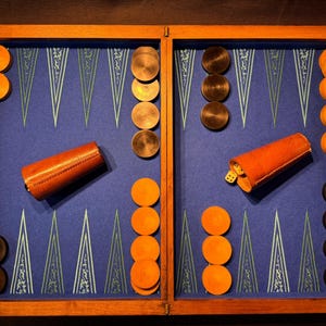 Puede incluir: Un tablero de backgammon de madera con una superficie de juego azul y un patrón decorativo. Incluye fichas naranjas y marrones, dos cubiletes de dados de cuero y dados. El tablero está abierto y listo para jugar.