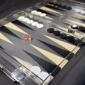 May include: A clear acrylic backgammon board with black and white triangular markings. The game board has white and black game pieces, and two red dice. The board is open, ready for play.
