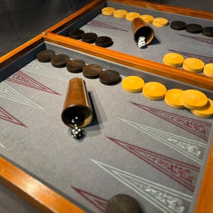 Op de afbeelding: Een houten backgammonbord met een grijs gevoerd speelveld. Het bord is open en toont twee sets zwarte speelstukken en twee sets bruine speelstukken. Er zijn ook twee dobbelstenen en twee bekers om te dobbelen.