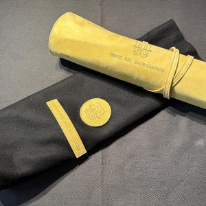 May include: A rolled-up, light yellow suede backgammon game case with a matching tie, resting on a black felt case. The text "SC GIOCO DEL BACKGAMMON" is visible on the suede case and a gold-colored emblem.
