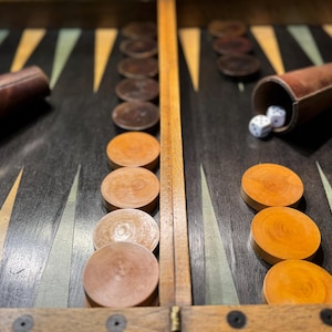 Può includere: Un tabellone di backgammon con pedine e dadi in legno. Il tabellone è scuro con segni triangolari in tonalità di giallo e verde. Ci sono coppette per dadi in pelle marrone su entrambi i lati del tabellone. Le pedine sono marroni e arancioni.