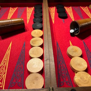 May include: A backgammon board with a red playing surface, wooden game pieces, and leather dice cups. The board features a geometric design in yellow and blue. Dice are visible in the cups and on the board.