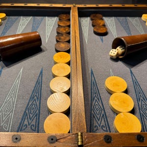 Può includere: Un gioco da tavolo del backgammon con pezzi e dadi di legno. Il tabellone è grigio con disegni geometrici blu e bianchi. Ci sono coppette per dadi in pelle marrone su entrambi i lati del tabellone. I pezzi del gioco sono in legno chiaro e scuro.