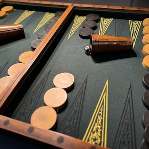 Può includere: Un tabellone da backgammon in legno con una superficie di gioco in feltro verde. Il tabellone è preparato per una partita con pedine nere e bianche. Ci sono due bicchieri da dado in pelle sul tabellone.