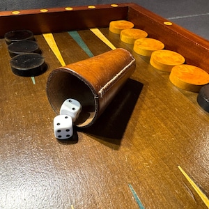 Può includere: Un tabellone di backgammon con struttura in legno, con un bicchiere per dadi in pelle marrone e due dadi bianchi. I pezzi del gioco sono neri e marroni. Il tabellone ha un design classico con accenti gialli e blu.