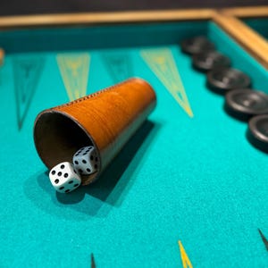 May include: A backgammon set with a teal playing surface. A brown leather dice cup is tilted, revealing two white dice with black dots. Black and tan game pieces are arranged on the board, ready for play. A classic board game.