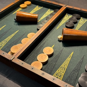 Può includere: Un set da backgammon in legno con un piano di gioco in feltro verde. Il set include due bicchieri da dado in pelle, due set di pedine e un dado bianco. Le pedine sono disposte a forma di un motivo sul tabellone.