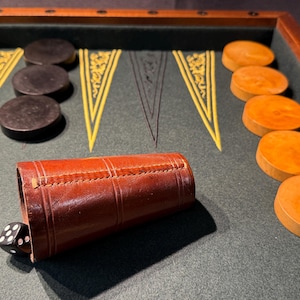 以下が含まれることがあります： 緑色のフェルトのボードに黒と茶色の駒が置かれたバックギャモンのゲームセット。駒は伝統的なバックギャモンのパターンで配置されています。手前に茶色の革製のサイコロカップがあります。