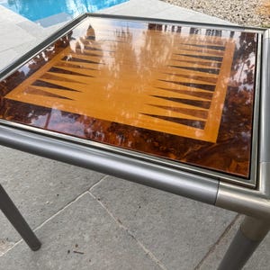 May include: A square game table featuring a backgammon board surface with a glossy finish. The board displays light wood triangles against a dark, marbled wood grain border. The table is supported by a silver-toned metal frame and four legs.