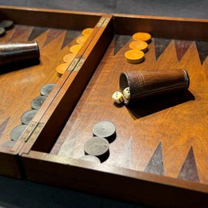 Puede incluir: Un tablero de backgammon de madera con fichas negras y blancas, dados de marfil y dos vasos marrones. El tablero está abierto y las piezas están esparcidas por el tablero.