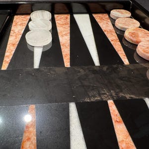 May include: A backgammon board with a black playing surface, featuring white and orange triangular points. White and orange game pieces are arranged on the board, ready for play. The board is made of a glossy material.