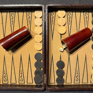 May include: Open backgammon board with wooden game pieces. The board is brown with a patterned interior. The game pieces are black and light brown. A dark red wooden dice cup is present, with dice inside.