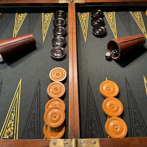Può includere: Un tabellone da gioco di backgammon in legno con una superficie di gioco in feltro verde. Il tabellone è aperto e mostra pedine nere e bianche, dadi e bicchieri. Il tabellone ha un design tradizionale con intagli intricati.