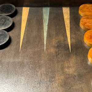 Può includere: Un tabellone da backgammon in legno con pedine nere e marroni. Il tabellone ha una superficie marrone scuro con tre triangoli dorati e blu.