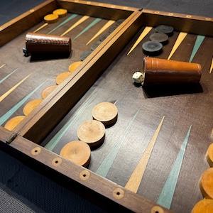 Può includere: Un set di backgammon vintage con cornice e superficie di gioco in legno. Il tabellone presenta triangoli chiari e scuri alternati, con pedine e coppette per dadi in legno. Il set include pedine e dadi in legno.