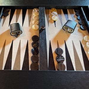 Può includere: Un tabellone di backgammon con una cornice in legno scuro e una superficie di gioco con triangoli chiari e scuri alternati. I pezzi sono di colori chiari e scuri e sono visibili dei dadi.