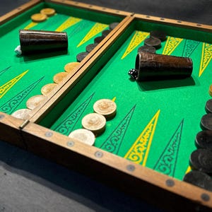 Può includere: Un tabellone di backgammon in legno con superficie di gioco verde e cornice in legno. Il tabellone presenta pedine marroni e beige, due coppette e dadi. La superficie verde ha elementi decorativi gialli e bianchi.