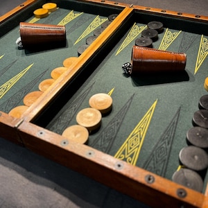 Può includere: Un tabellone di backgammon in legno con una superficie di gioco verde e disegni geometrici gialli. Il tabellone presenta coppette per dadi in pelle marrone, pedine in legno di colori chiari e scuri e dadi.