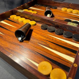 Könnte beinhalten: Ein Backgammon-Set aus Holz mit dunkler Maserung. Das Spielbrett zeigt abwechselnd schwarze und weiße Dreiecke, mit gelben und schwarzen Spielsteinen. Würfelbecher und Würfel sind ebenfalls vorhanden, spielbereit.