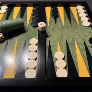 Può includere: Un tabellone di backgammon con una superficie di gioco verde e gialla, cornice nera e pedine avorio e nere. Sono presenti un bicchiere per dadi e dadi. I dadi mostrano i numeri 64 e 16.