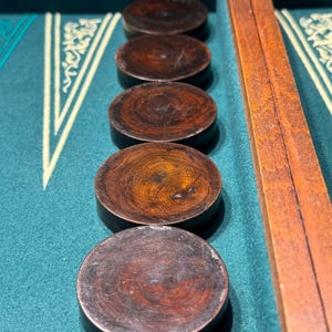 Può includere: Primo piano di una tavola da backgammon con pedine in legno marrone scuro. Le pedine circolari sono disposte in fila su una superficie di feltro verde. Un telaio in legno è visibile sul lato destro.