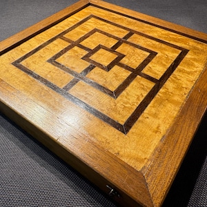 May include: A square wooden game board featuring a Nine Men's Morris pattern inlaid with dark brown wood against a lighter, figured golden-brown wood surface. The board is framed by a thick, solid wood border with visible grain and metal latches.