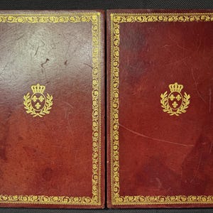 May include: Two antique books with burgundy leather covers. Each book features a gold-colored decorative border and a gold emblem of a crown and wreath. The books are side-by-side, suggesting a set.