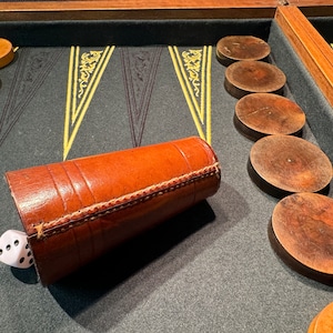 Puede incluir: Un vaso de dados de cuero marrón con un dado blanco sobre un tablero de backgammon de fieltro verde con fichas de madera.