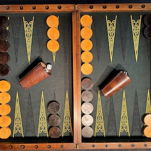 Può includere: Un set da gioco di backgammon in legno con un piano di gioco in feltro verde. I pezzi del gioco sono disposti sulla scacchiera e ci sono due bicchieri da dado in pelle.