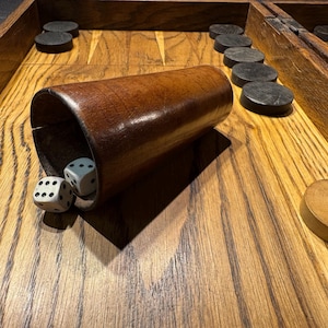 May include: A backgammon set with a wooden board and pieces. A brown wooden dice cup is tipped over, revealing two dice. The game pieces are black and tan. The board is made of wood with a natural grain.