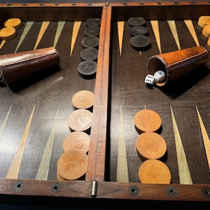 Può includere: Un tabellone da backgammon in legno con pedine nere e bianche, bicchieri da dado in pelle marrone e dadi bianchi. Il tabellone è aperto e pronto per giocare.