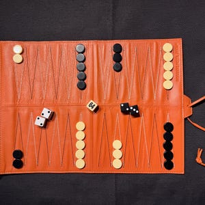 May include: An orange leather backgammon set, rolled out on a dark surface. The set includes black and cream-colored game pieces, dice, and a die with the number 64. The set is rolled up with a zipper closure and a leather tie.