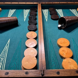 Può includere: Un tabellone di backgammon con una superficie di gioco verde acqua, cornice in legno e coppette per dadi in pelle marrone. I pezzi del gioco sono in legno e neri. Un dado bianco è visibile.