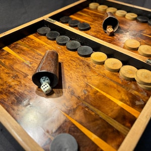 Könnte beinhalten: Ein Vintage-Backgammon-Set mit einer Holzspieloberfläche und einem Holzrahmen. Das Set enthält schwarze und beige Spielsteine, zwei Würfelbecher und Würfel. Die Spieloberfläche hat ein warmes Holzmaserungsmuster mit gelben Akzenten.