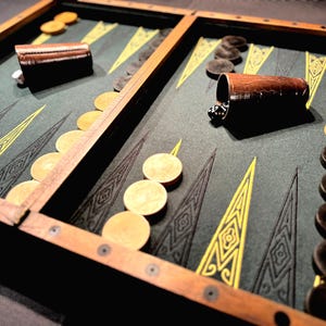 Può includere: Un tabellone da gioco di backgammon in legno con una superficie di gioco in feltro verde. Il tabellone è aperto e mostra le pedine nelle loro posizioni di partenza. Le pedine sono in legno e sono dipinte di nero e bianco.
