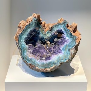 May include: A large, open geode with a rough, brown exterior and a vibrant interior. The interior features layers of light blue, teal, and deep purple crystals. The geode is displayed on a white pedestal.