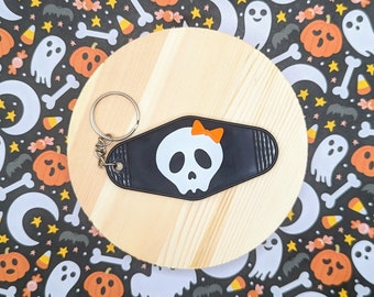 May include: A black and white key chain with a skull and crossbones design. The skull has a pink bow on top. The key chain is on a wooden surface with a Halloween theme.