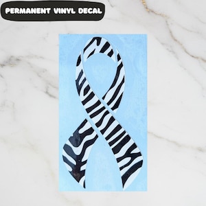 May include: A permanent vinyl decal featuring a ribbon design with a zebra print pattern in black and white. The ribbon is set against a light blue background. The text "PERMANENT VINYL DECAL" is displayed in a black banner at the top.