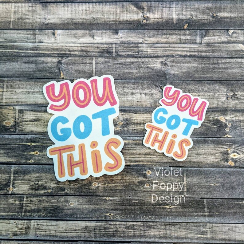 May include: Two stickers with the text "You Got This" in different sizes. The larger sticker has a pink, orange, and blue color scheme. The smaller sticker has a blue, orange, and white color scheme.
