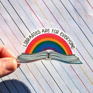 May include: A sticker featuring a rainbow arching over an open book. The text "Libraries are for everyone" is printed in black. The sticker has a white border and is held against a background of colorful diagonal stripes.